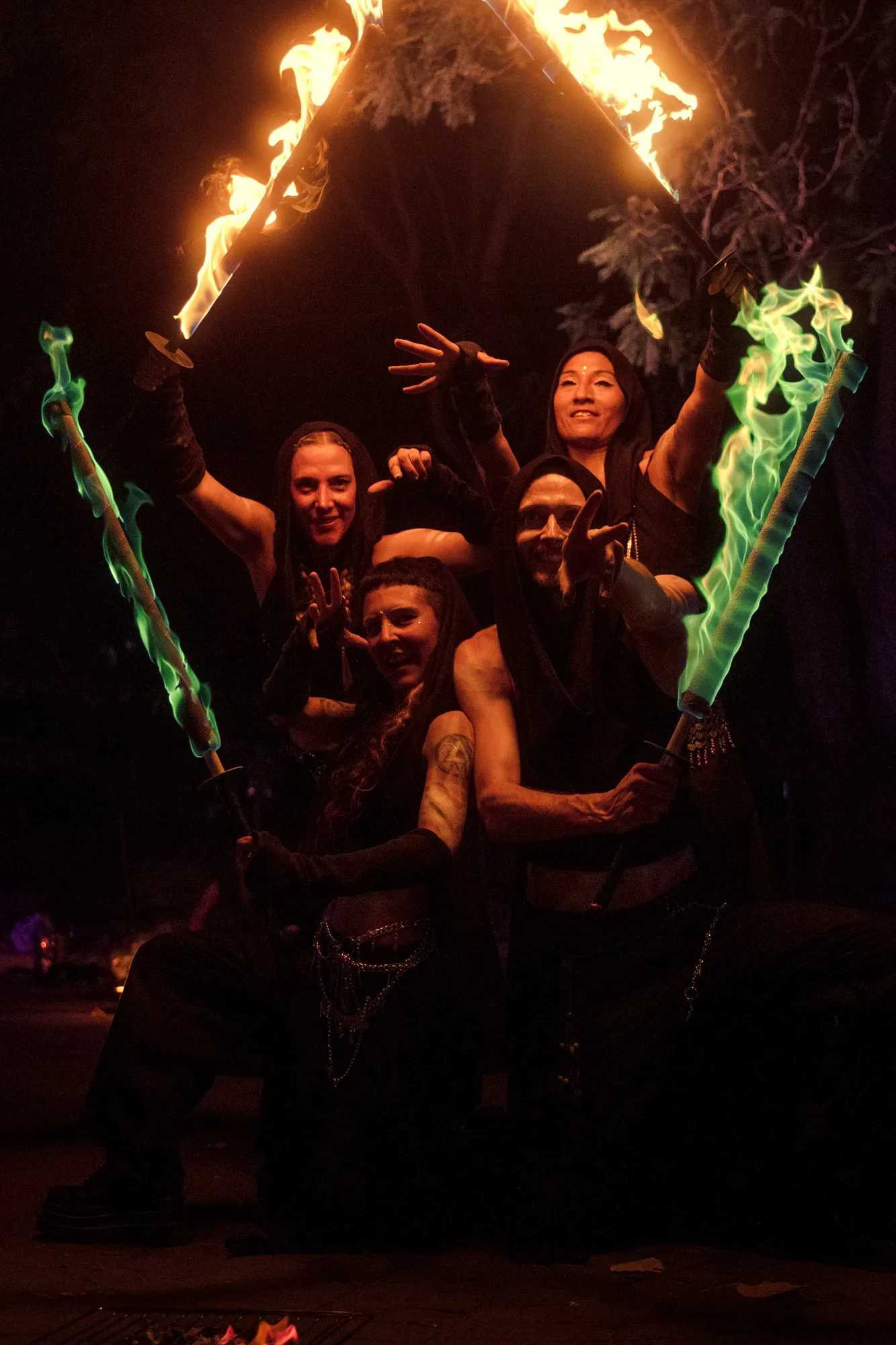 Four performers in black hooded costumes hold flaming swords outdoors at night — orange flames raised above, green flames below
