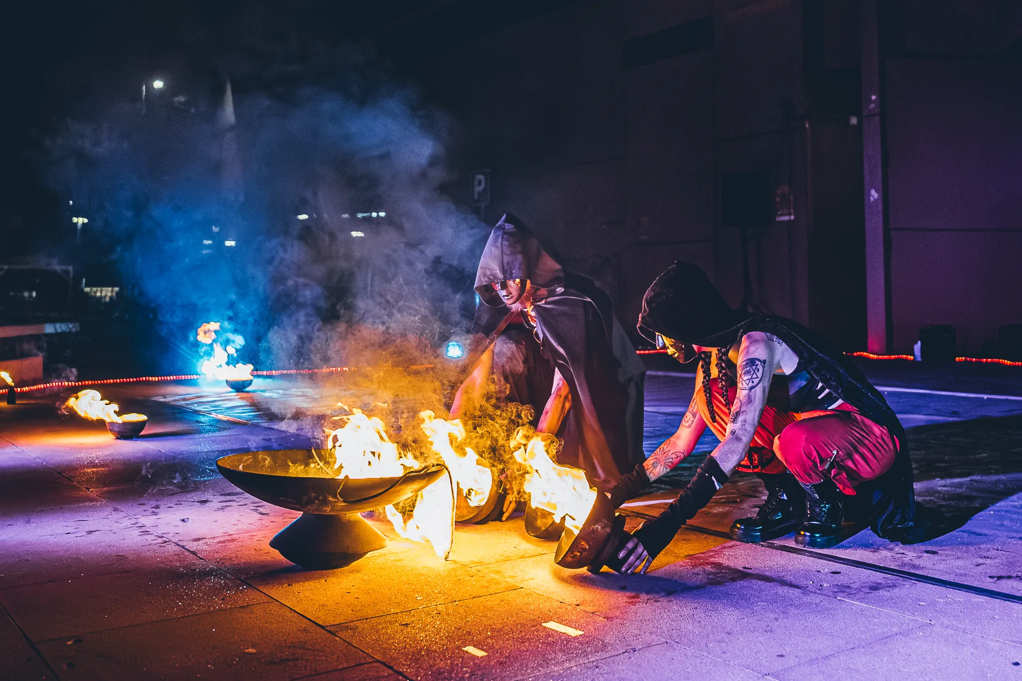 Two hooded performers crouch at night with flaming fire bowls and thick smoke rising around them
