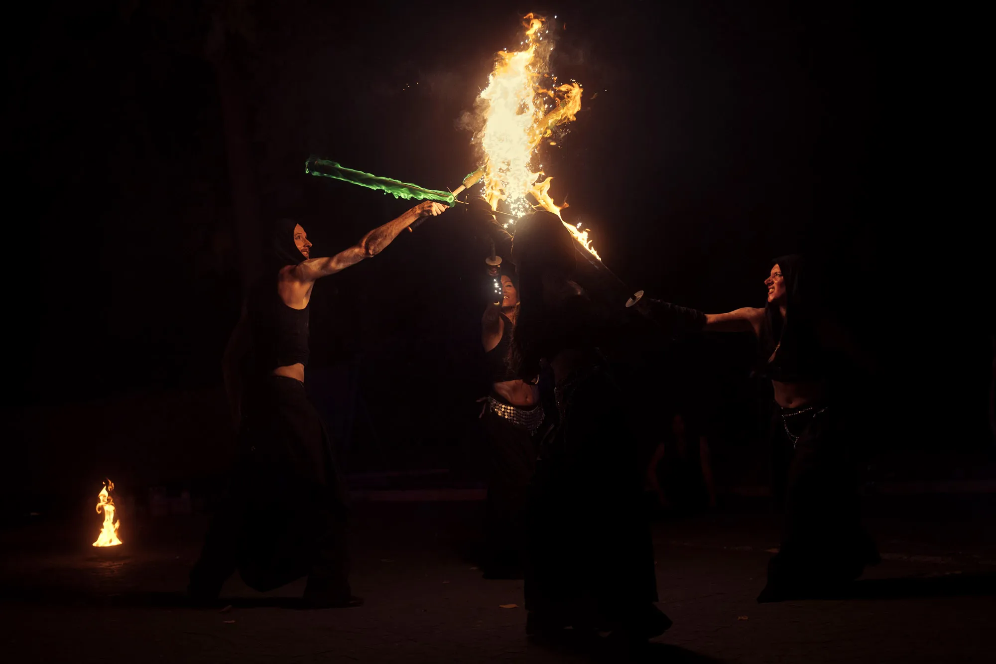 Three performers in dark clothing at night; a central staff erupts in orange flame and sparks while another holds a glowing green staff, with a small flame on the ground