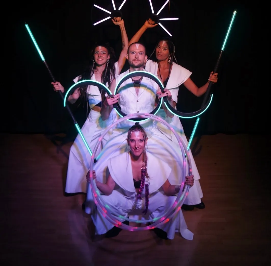 Four performers in white costumes posing with glowing LED fans, staffs, and hoops against a dark background